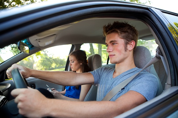 A teenage boy in a short sleeved t-shirt steers the wheel of a car while a young female passenger looks down at her phone.
