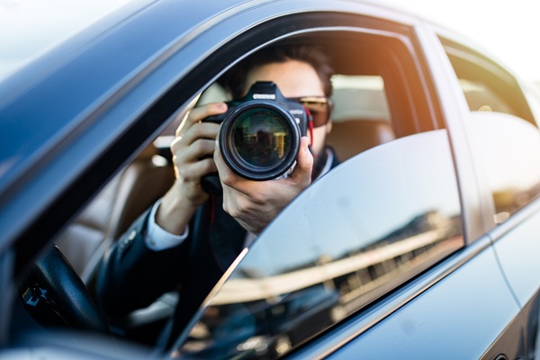 A man sits in the driver's seat of a car, partially obscured by the window, holding a professional DSLR camera up to his eye to take a photo for an insurance company.