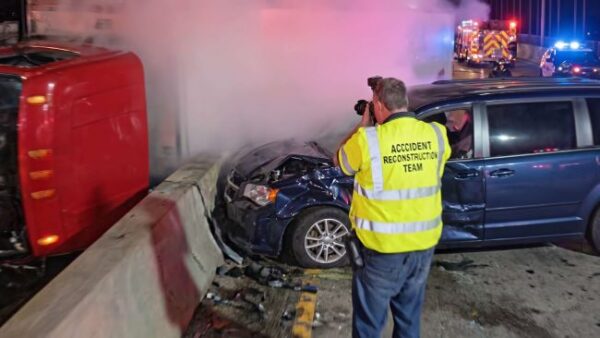 Accident reconstruction team member photographing a Cleveland truck accident, with a heavily damaged blue minivan beside an overturned red semi-truck on a highway at night, and emergency vehicles in the background.
