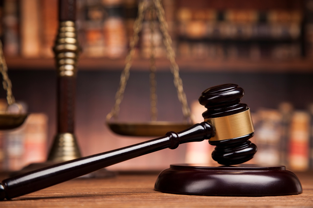 A close-up of a dark wooden judicial gavel resting on its sound block on a wooden table, with the blurred scales of justice and law bookshelves in the background.